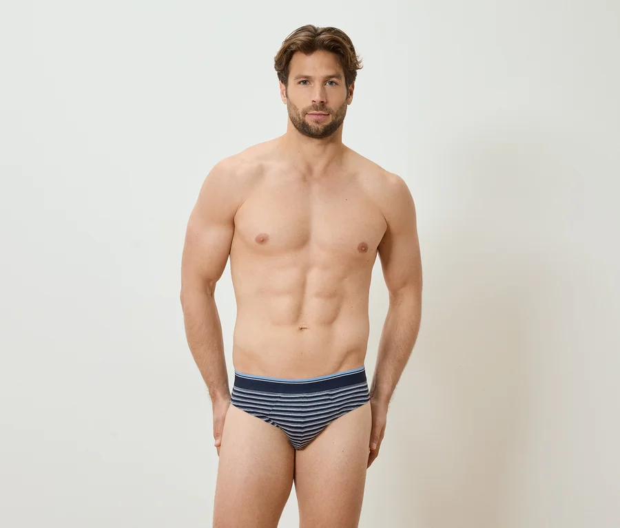 Mann mit freiem Oberkörper und gestreifter Unterhose steht vor einer weißen Wand.