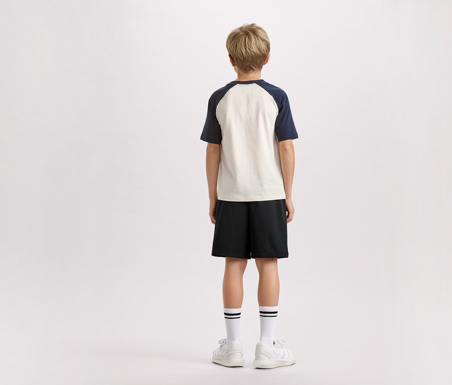 Rückansicht eines blonden Kindes mit T-Shirt mit marineblauen Ärmeln, schwarzen Shorts, Socken mit Streifen und weißen Turnschuhen vor einem weißen Hintergrund.