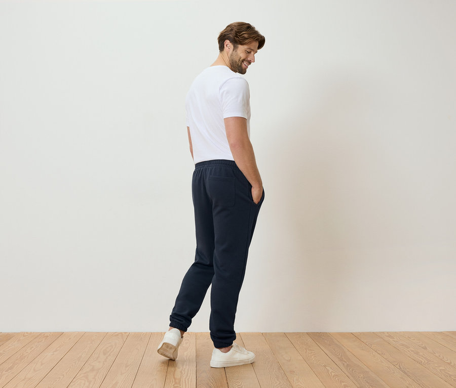 Mann in weißem T-Shirt und blauer Jogginghose steht auf einem Holzboden.