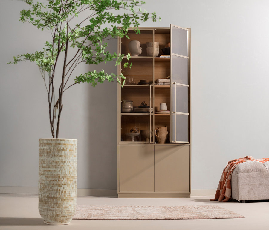 Einrichtungsszene mit einem hohen beigen Schrank mit teilweise geöffneter Glastür, einem beigen Hocker mit einer orangefarbenen Decke darauf, einem gemusterten Teppich und einer hohen beigen Vase mit einem grünen Baum darin.