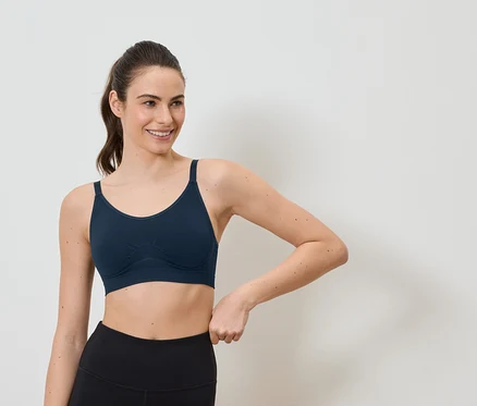 Lächelnde Frau mit Pferdeschwanz trägt einen marineblauen Sport-BH und schwarze Leggings vor einem weißen Hintergrund.