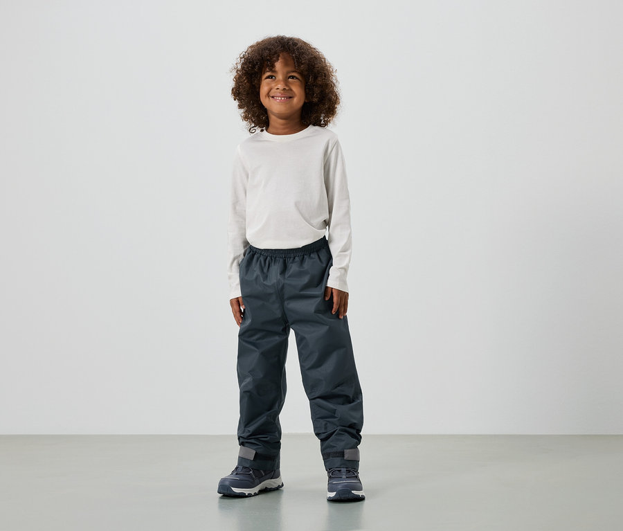Ein lächelnder Junge mit lockigem Haar trägt ein langärmliges Oberteil, Regenhose und Turnschuhe.