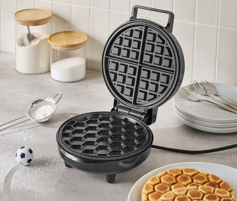 Offenes, schwarzes Waffeleisen auf einer Küchentheke mit fertigen Waffeln auf einem Teller, einem kleinen Fußball und Zutaten in Gläsern im Hintergrund.