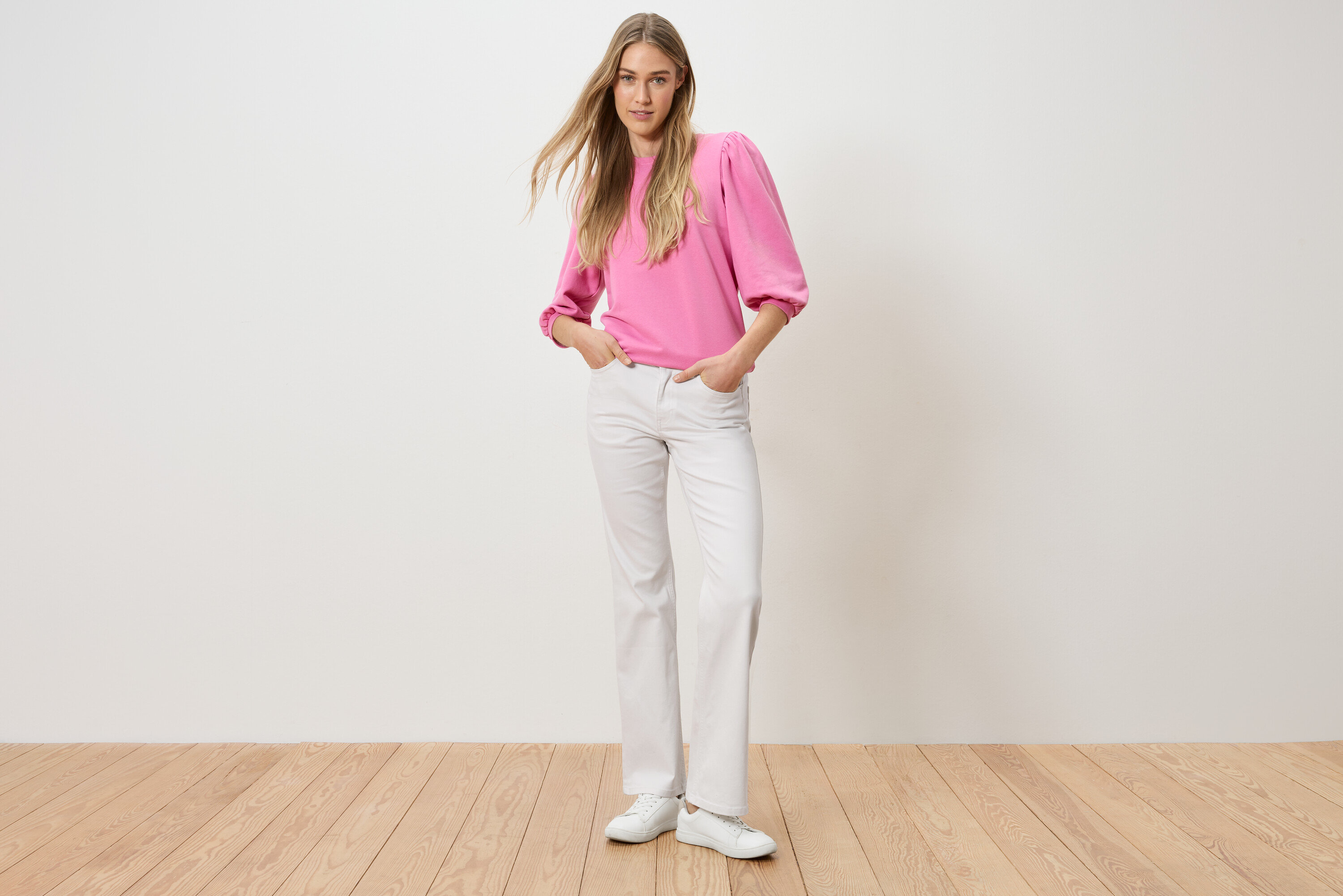 Eine Frau steht mit ihren Händen in den Hosentaschen. Sie trägt ein pinkes Oberteil und weiße Jeans.