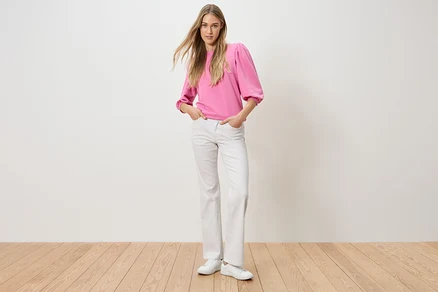 Eine Frau steht mit ihren Händen in den Hosentaschen. Sie trägt ein pinkes Oberteil und weiße Jeans.