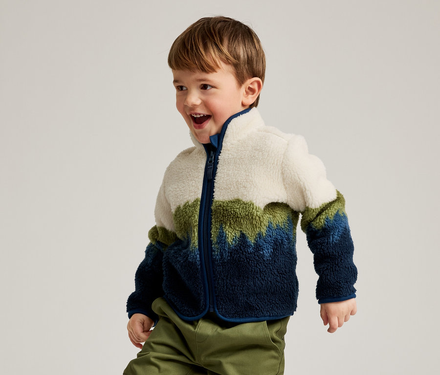 Ein kleiner Junge mit braunen Haaren, der eine Fleecejacke in Weiß, Grün und Blau sowie grüne Hosen trägt, lächelt und blickt nach rechts.