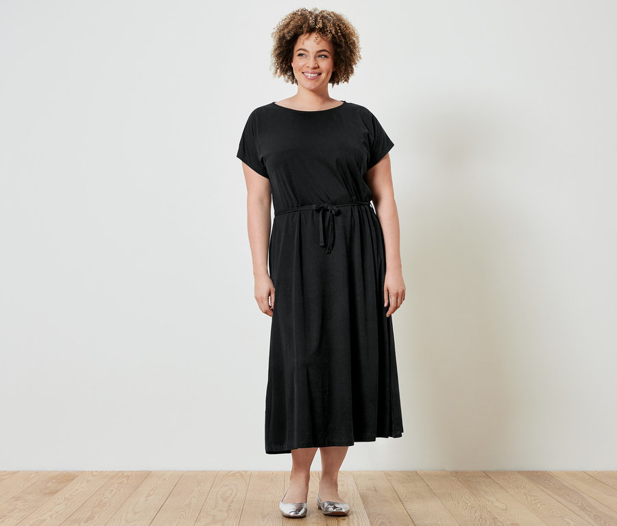 Frau mit lockigem Haar trägt ein schwarzes Kleid mit Kordelzug und silberne Ballerinas vor einer weißen Wand.