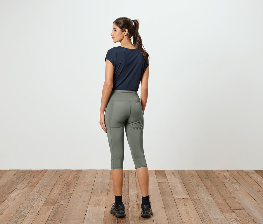 Frau mit Pferdeschwanz, von hinten gesehen, trägt ein marineblaues T-Shirt und olivgrüne Capri-Leggings vor einer weißen Wand.