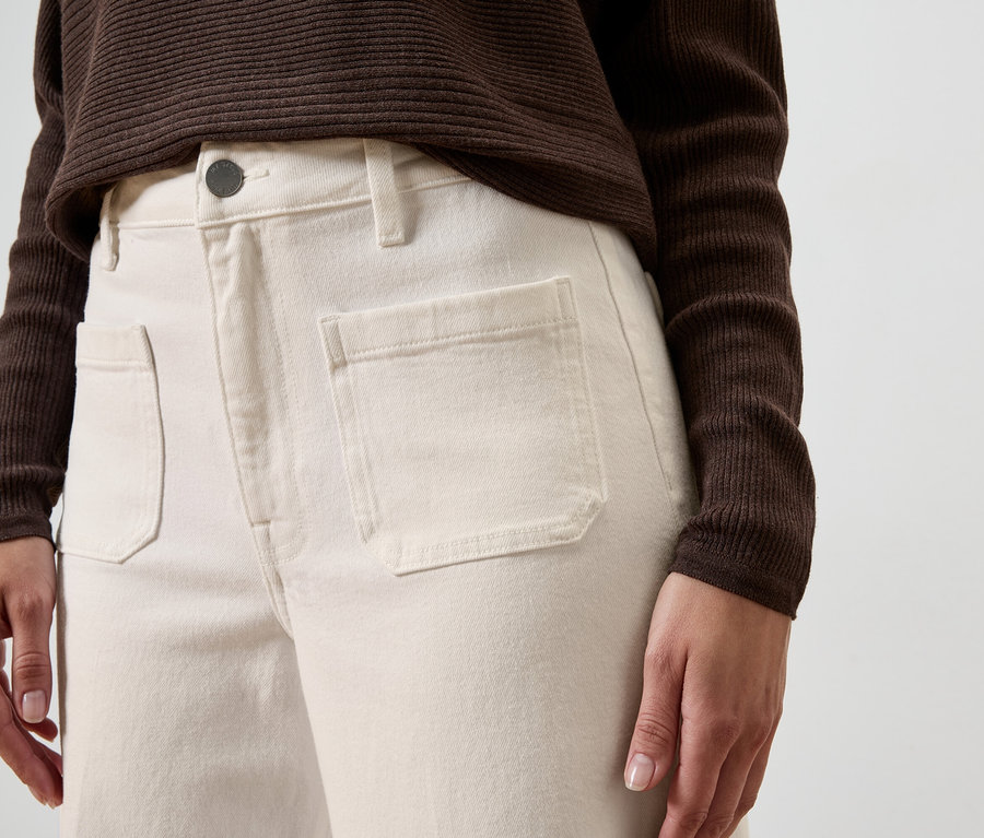 Nahaufnahme einer Person in cremefarbenen Hosen mit aufgesetzten Taschen und einem braunen Oberteil vor einem neutralen Hintergrund.