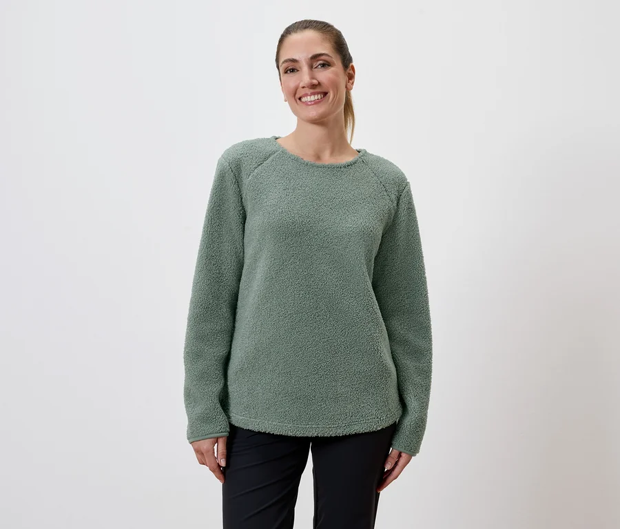 Eine lächelnde Frau mit Pferdeschwanz trägt einen grünen Fleecepullover und schwarze Hosen vor einem weißen Hintergrund.