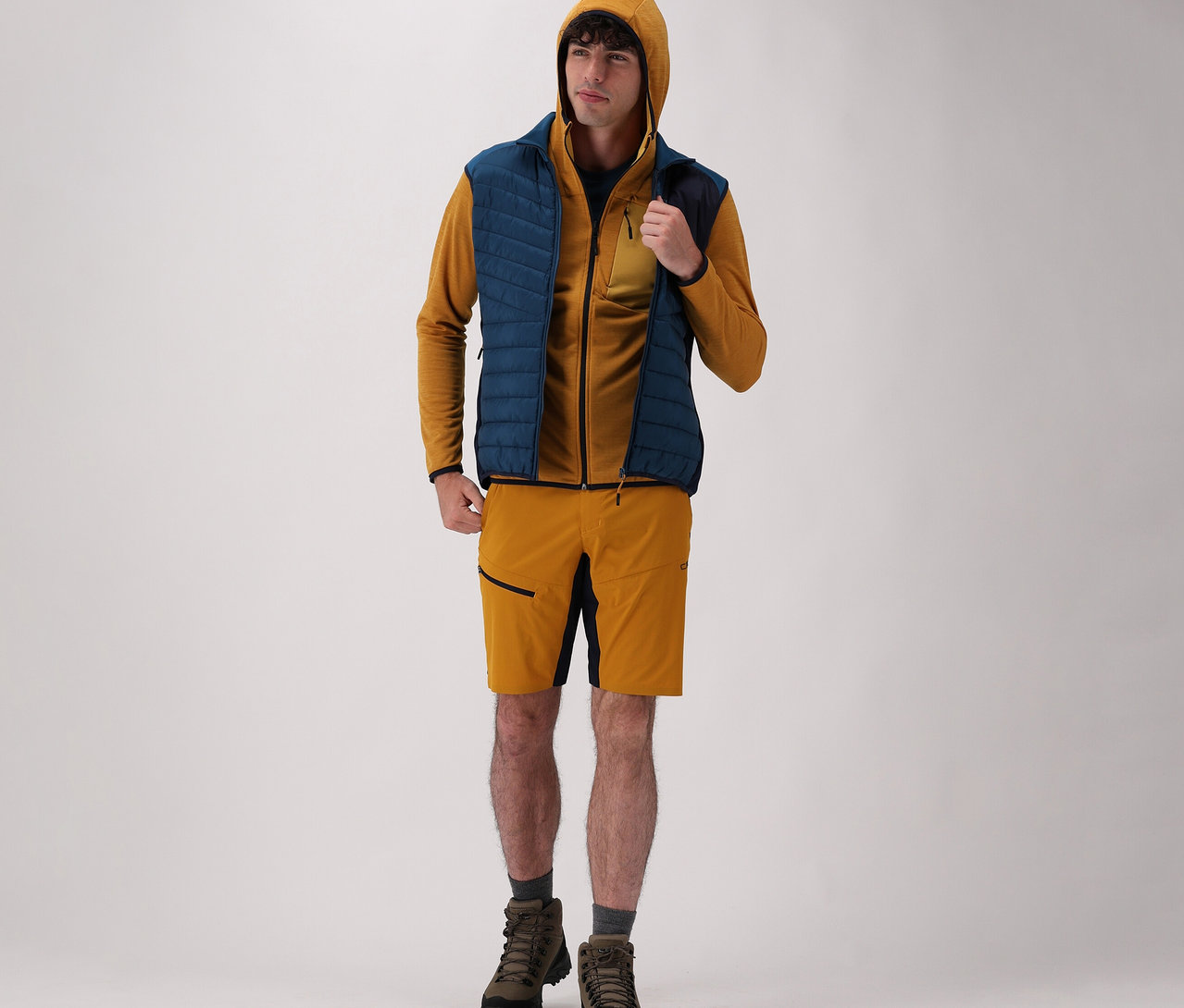 Mann trägt CMP Herren-Kapuzen-Jacke, CMP Herren-Hybridweste, Shorts und Stiefel vor einem hellen Hintergrund.