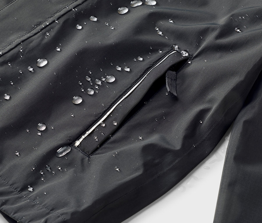 Nahaufnahme einer schwarzen Jacke mit wasserabweisender Oberfläche und einer Reißverschlusstasche mit weißem Rand, auf der Wassertropfen zu sehen sind.