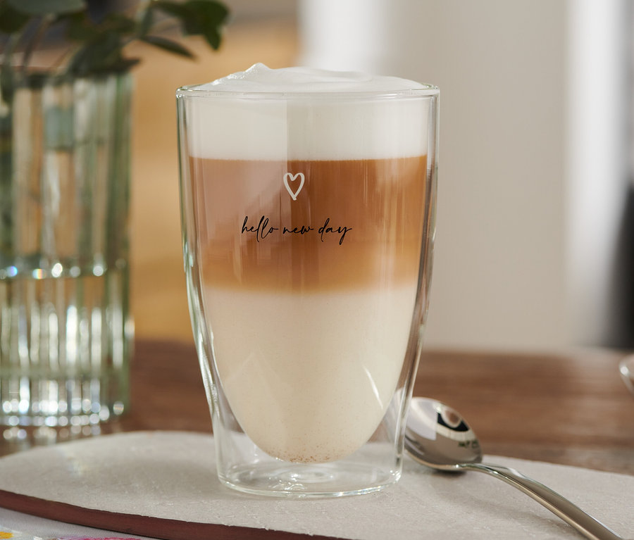 Ein Latte Macchiato Glas, bedruckt mit Herzchen, gefüllt mit Milch-, Kaffee- und Schaumschichten.