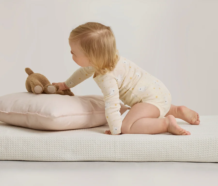 Ein Baby in einem gemusterten Body kniet auf einer weißen, strukturierten Oberfläche und greift nach einem braunen Teddybär, der auf einem rosa Kissen liegt.