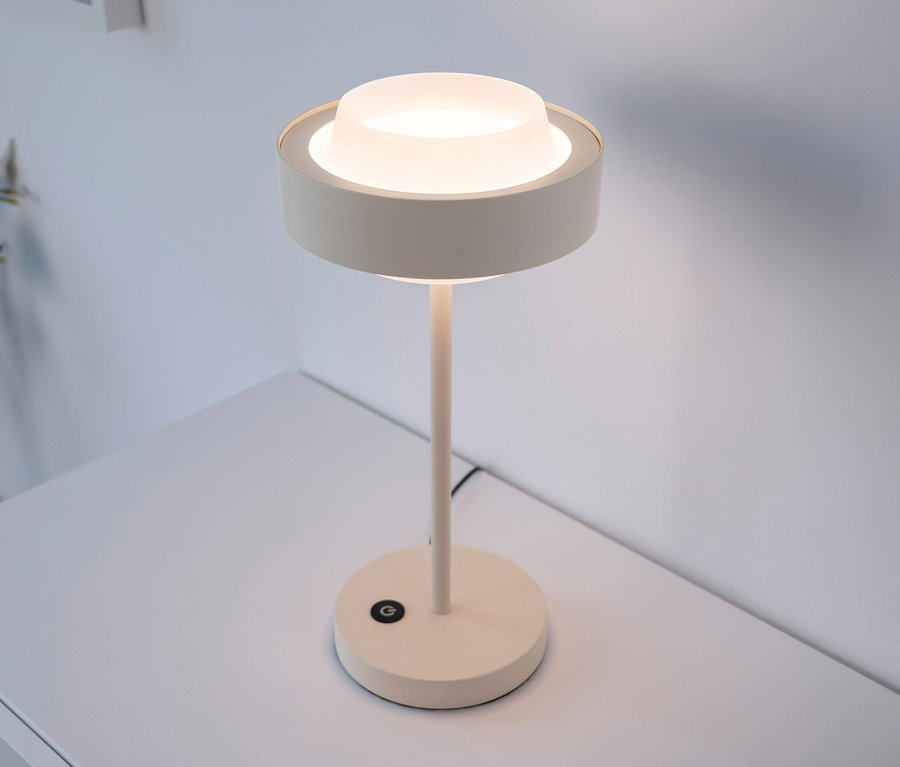 Eine moderne, cremefarbene Tischlampe mit einem runden, mattierten Diffusor leuchtet sanft auf einem weißen Schreibtisch vor einer weißen Wand.
