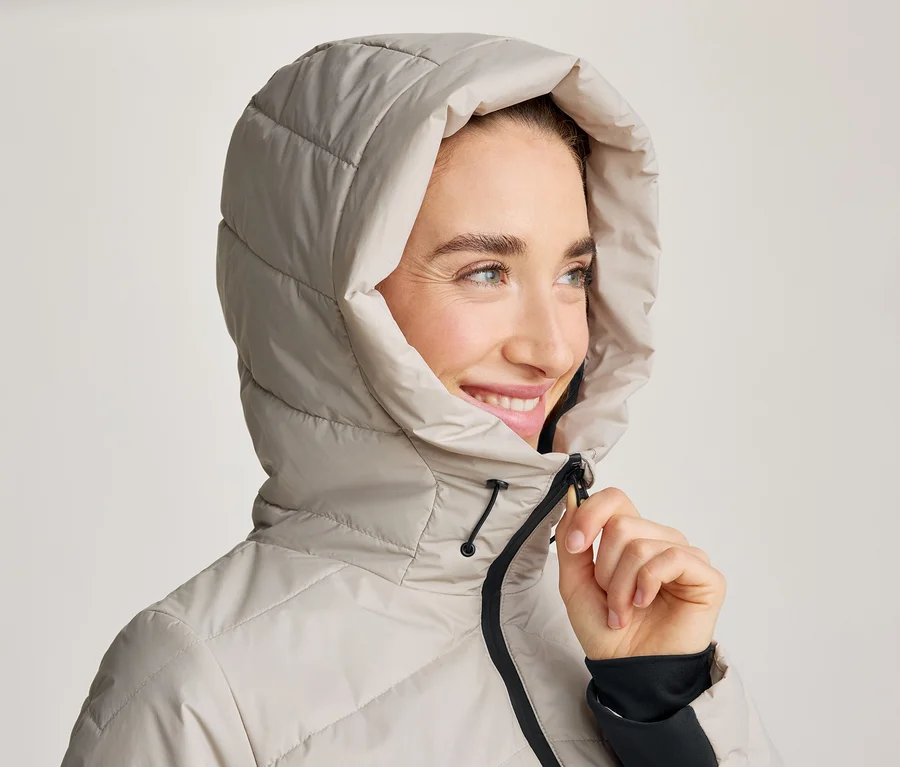 Eine lächelnde Frau trägt eine hellbeige Kapuzen-Steppjacke und blickt nach rechts. Ihre Hand ist am Reißverschluss der Jacke.