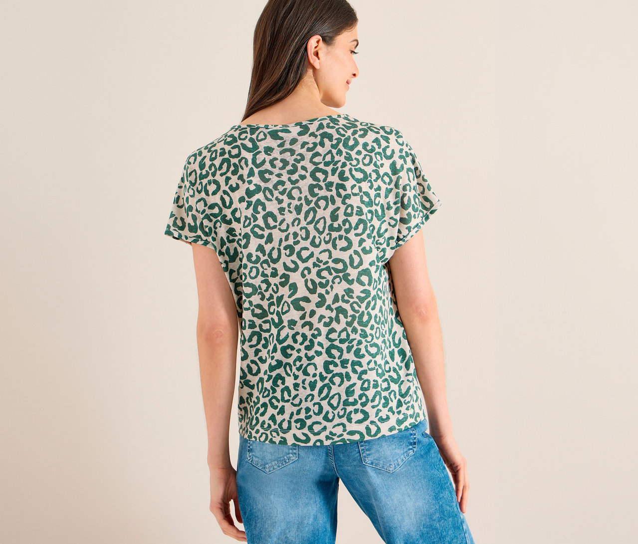Eine Frau von hinten, die ein beiges CECIL Kurzarmshirt mit grünem Leopardenmuster und blaue Jeans trägt. Sie blickt nach rechts.