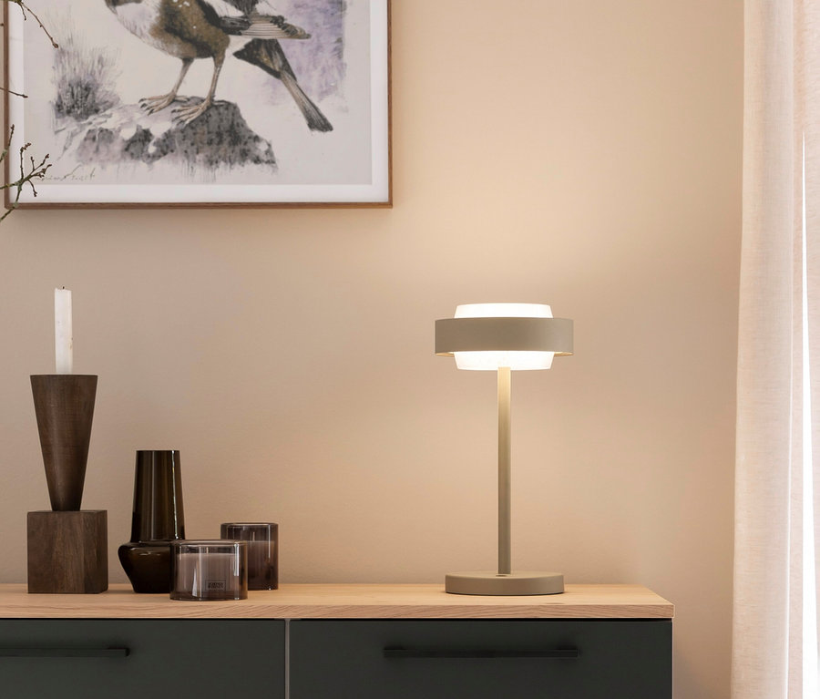 Eine moderne graue Tischlampe mit leuchtendem Licht steht auf einer Holzschrankplatte neben einem dunklen Kerzenhalter aus Holz, einer Vase und zwei dunklen Glaskerzenhaltern. Ein gerahmter Vogelkunstdruck hängt an der beigen Wand darüber.