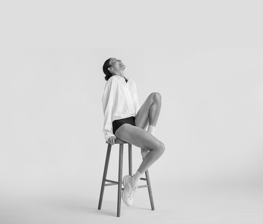 Frau sitzt lächelnd auf einem Hocker, trägt eine weiße Lange Leinenbluse, schwarze Unterwäsche, weiße Socken und weiße Sneaker.