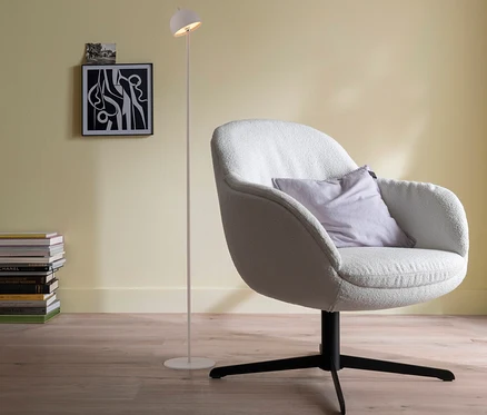 Ein weißer Sessel mit lila Kissen steht auf Holzboden neben einer weißen SCHÖNER WOHNEN-Kollektion Akku-Stehleuchte »Bellis«, Sandbrise. Links sind ein Bücherstapel und ein abstraktes Bild an der hellgelben Wand.