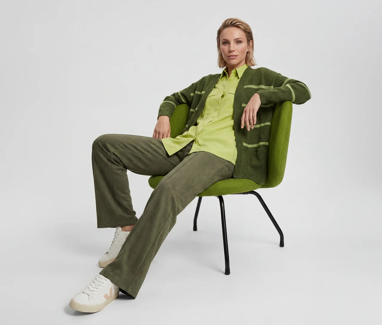 Frau mit kurzen blonden Haaren sitzt auf einem grünen Stuhl und trägt eine dunkelgrüne Strickjacke mit hellgrünen Streifen, ein hellgrünes Hemd, dunkelgrüne Hosen und weiße Sneaker.