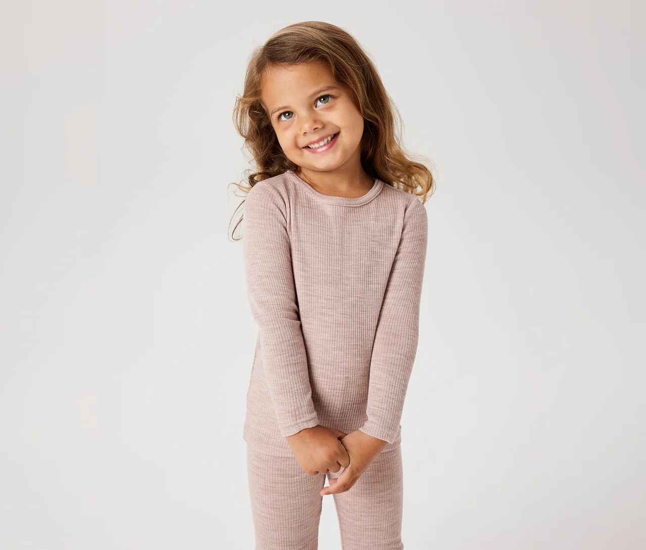 Lächelndes kleines Mädchen mit langen braunen Haaren, das ein geripptes, hellrosa Langarmshirt und passende Hose trägt, die Hände gefaltet.