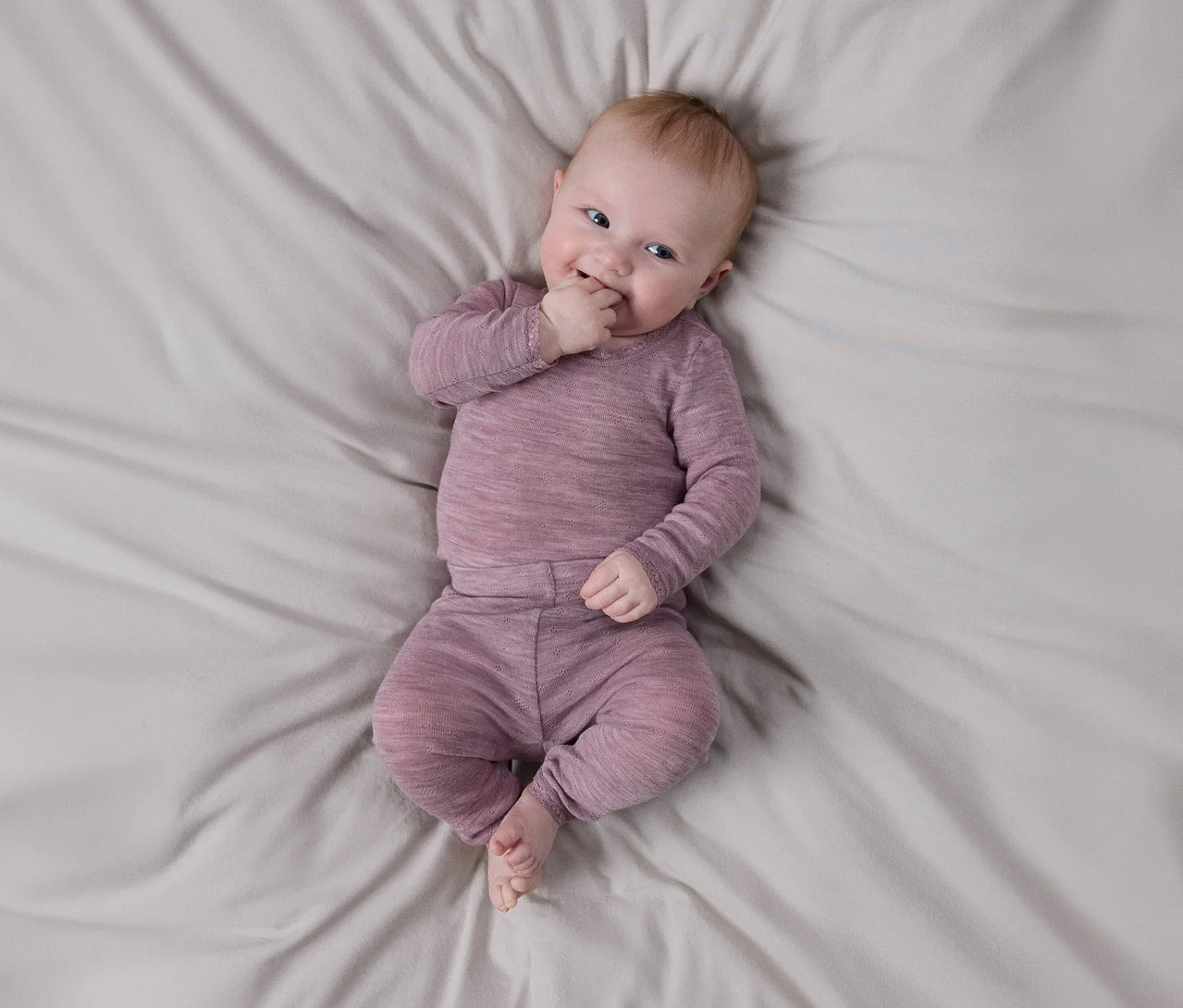 Lächelndes Baby liegt auf hellgrauem Laken, trägt langärmeliges, strukturiertes lila Oberteil und passende Hose, eine Hand am Mund.