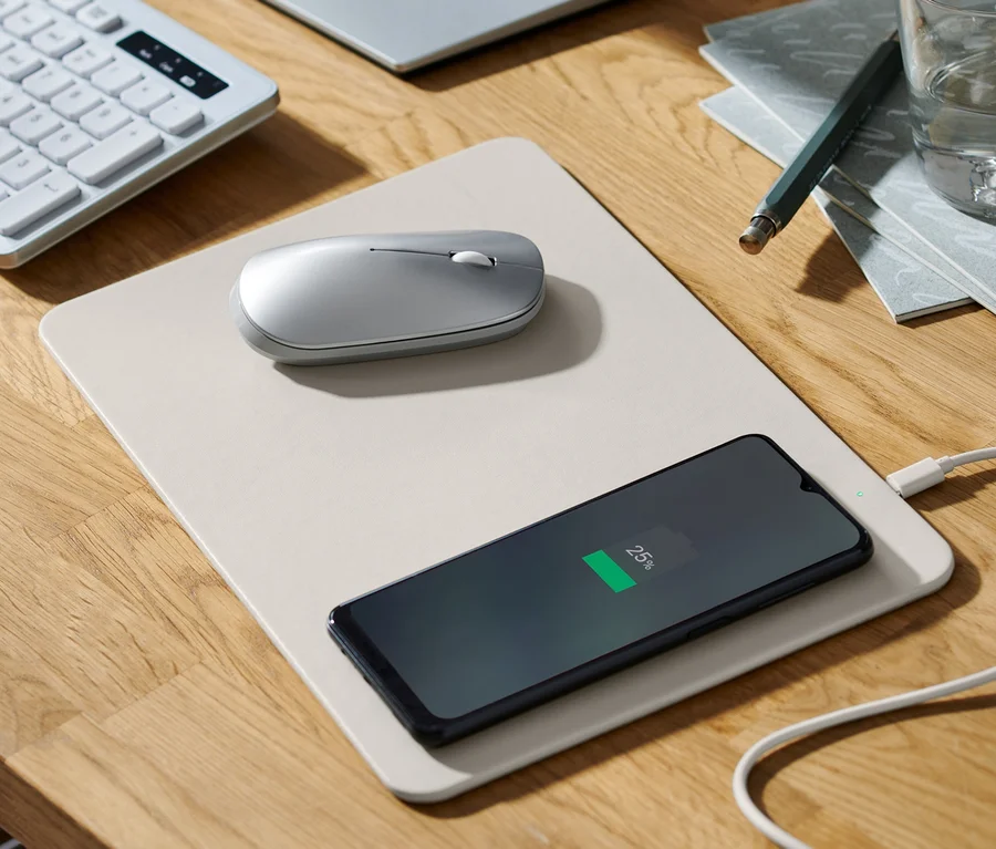 Auf einem Holztisch liegen ein Mauspad mit integriertem Ladegerät, auf dem ein Handy geladen wird, eine Maus, eine Tastatur, ein Monitor, ein Glas Wasser und ein Notizblock.
