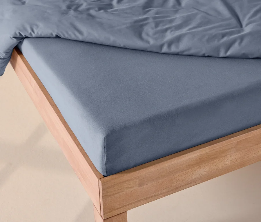 Bett mit blauem Jersey-Spannbettlaken und einer blauen Bettdecke auf einem hellen Holzrahmen.