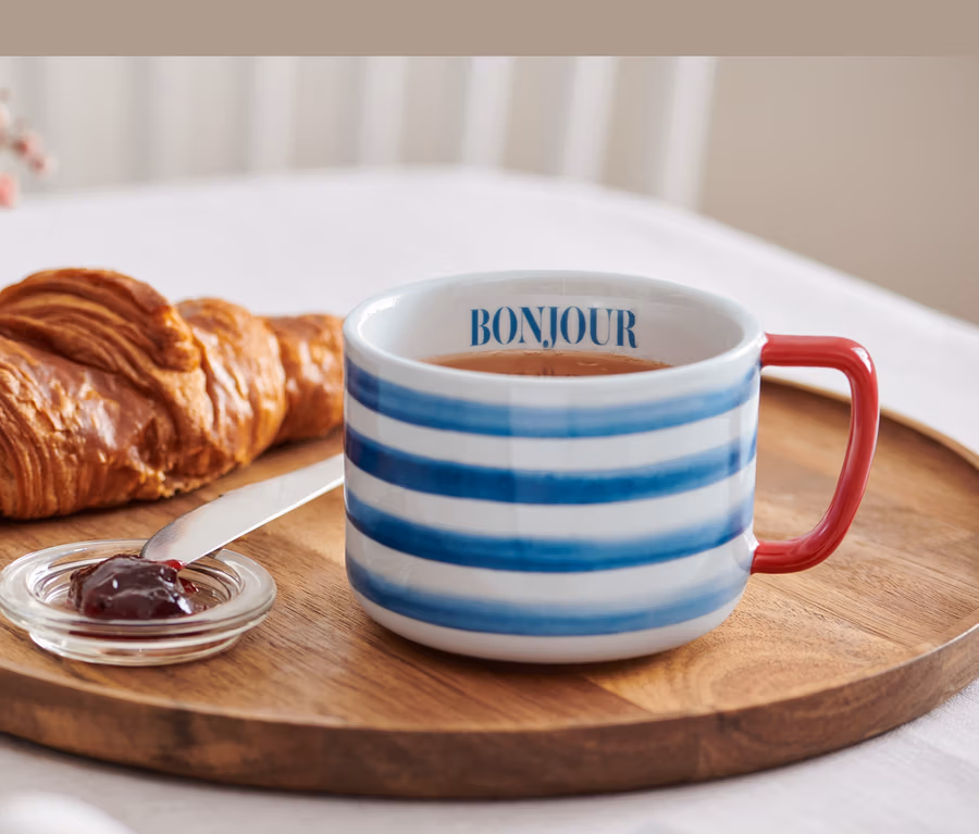 Auf einem Holztablett liegen ein Croissant, Marmelade und eine Jumbotasse mit der Aufschrift Bonjour.