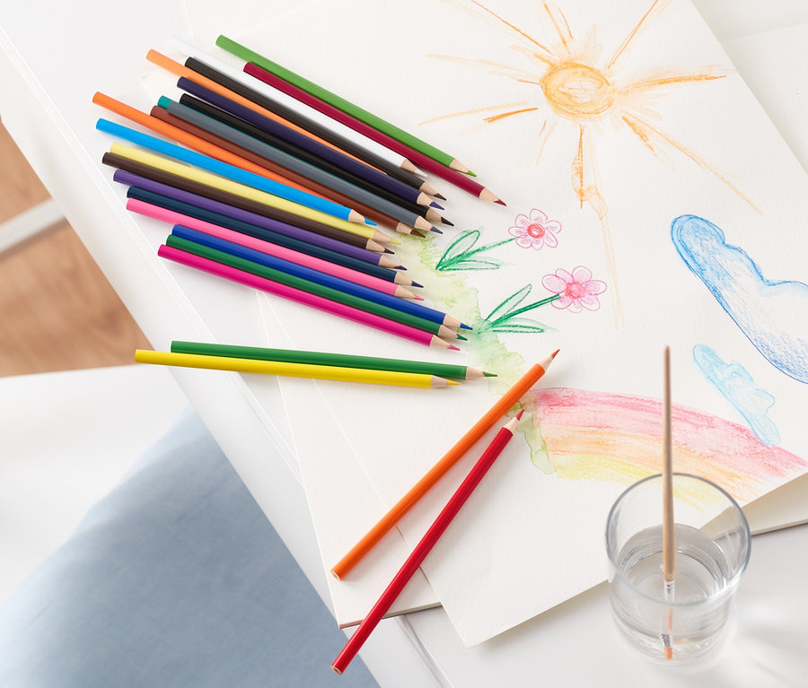 Auf einem weißen Tisch liegen eine Zeichnung von Sonne, Blumen, Wolken und einem Regenbogen, daneben Aquarell-Buntstifte und ein Glas mit Wasser und Pinsel.