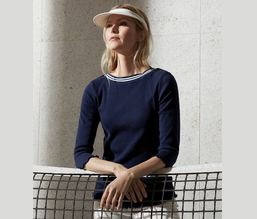 Blonde Frau mit Schirmmütze und Shirt mit 3/4-Arm lehnt an einem Tennisnetz.