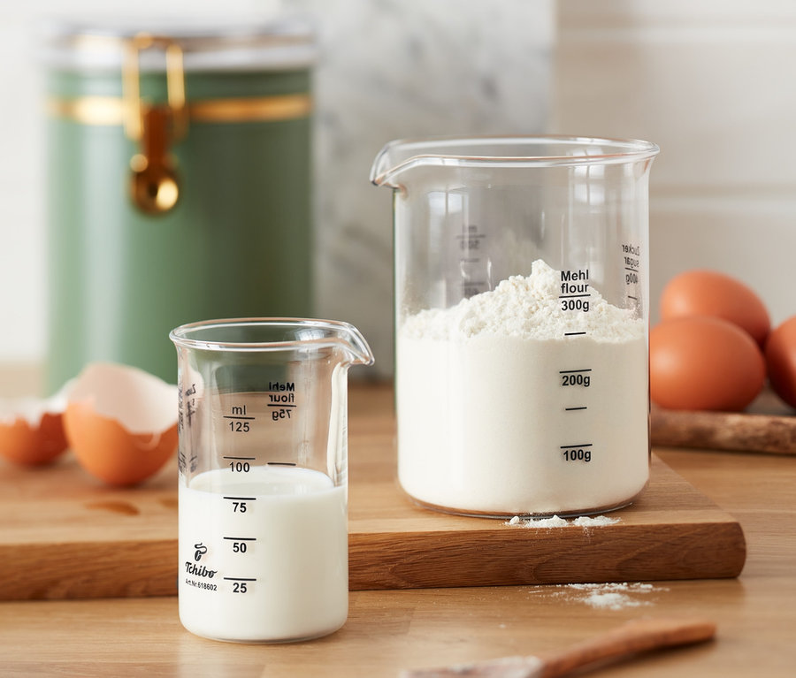 Zwei Messbecher, einer mit Milch und einer mit Mehl, stehen auf einem Holzbrett neben Eiern.