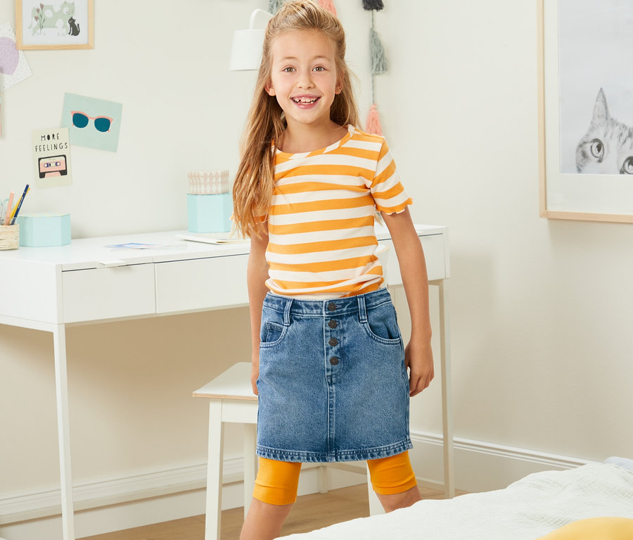 Lächelndes Mädchen posiert in Jeansrock vor einem Schreibtisch.