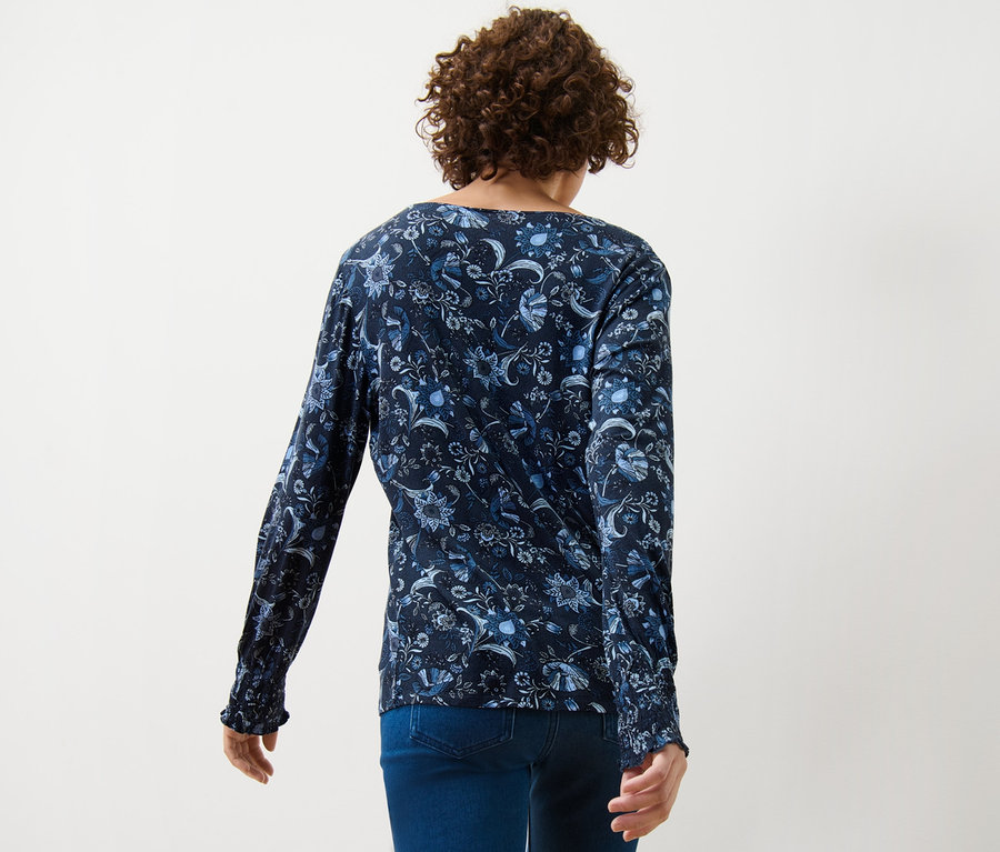 Rückansicht einer Frau in einem bedruckten Blusenshirt und blauen Jeans.