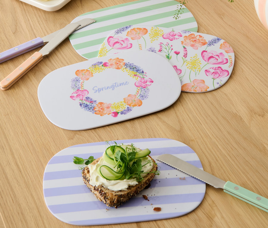 Vier Frühstücksbretter mit verschiedenen Mustern, Buttermesser und Brot mit Käse und Gurke auf einem Holztisch.