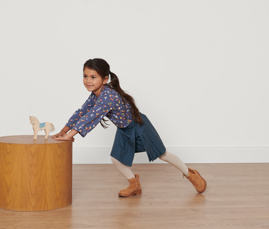 Mädchen lehnt sich an Tisch mit Holzpferd in blauem Blumenhemd und dunkelblauem Kleinkinder-Kiltrock.