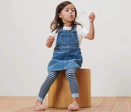 Mädchen sitzt auf einem Holzhocker und macht Seifenblasen. Sie trägt ein Jeanskleid und 2 Kinder-Feinstrumpf-Leggings.