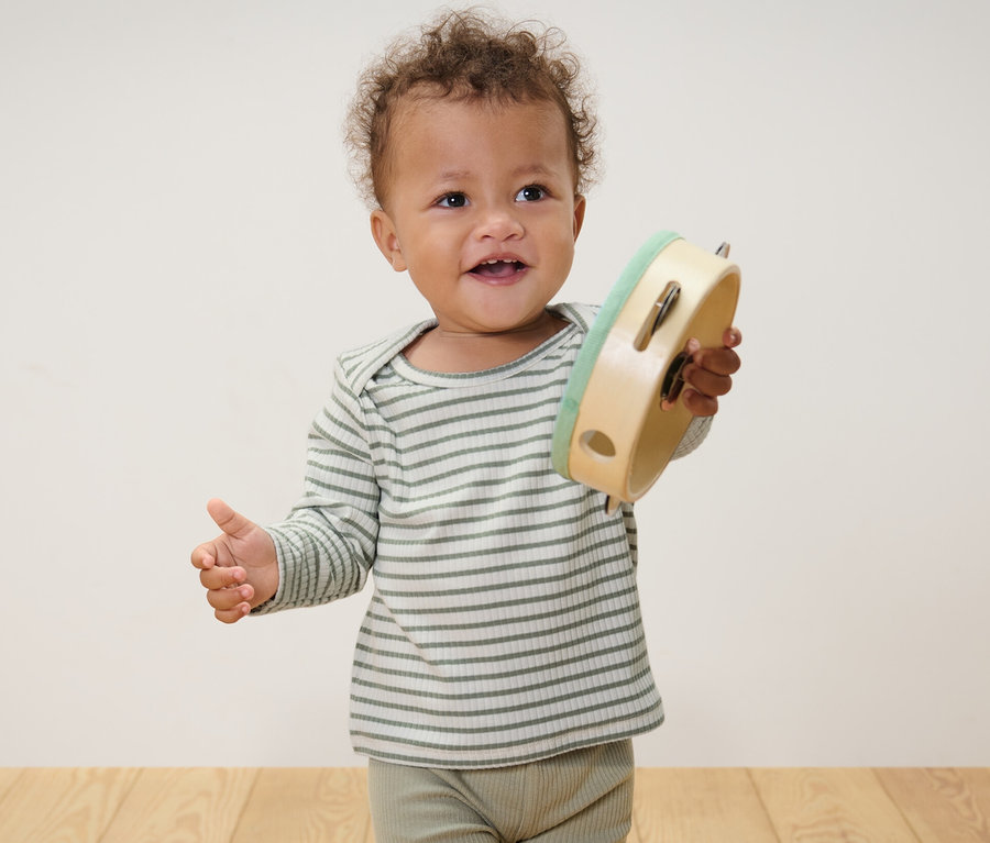 Ein Baby steht und hält ein Tamburin. Es trägt hellblaue und grüne Baby-Langarmshirts mit Rippstruktur.