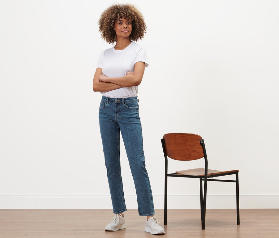 Eine Frau mit verschränkten Armen steht neben einem Stuhl und trägt eine Straight-Denim »Fit Juna« in Midblue.