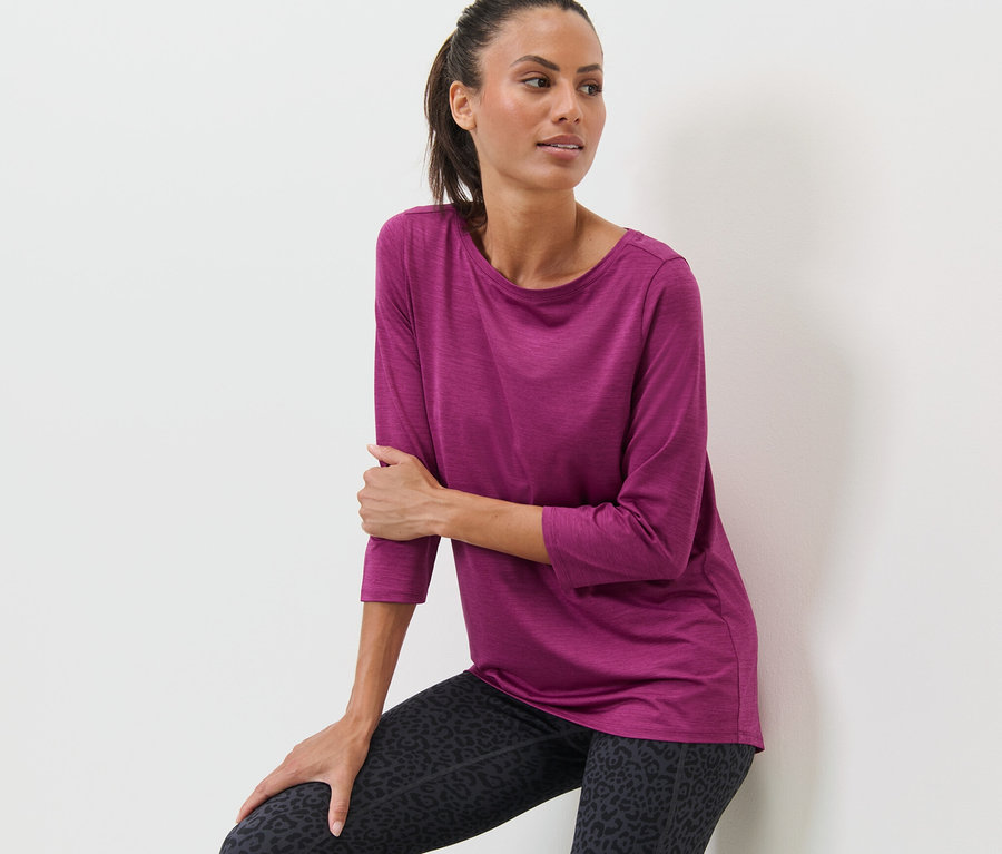 Eine Frau, die sich an eine Wand lehnt, trägt ein pinkes Funktionsshirt mit 3/4-Arm und Leggings mit Animal-Print.