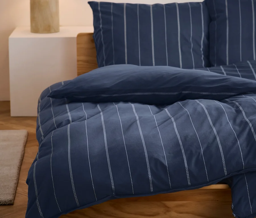 Blau gestreifte Winterjersey-Bettwäsche liegt auf einem Bett. Auf einem Nachttisch steht eine Lampe.