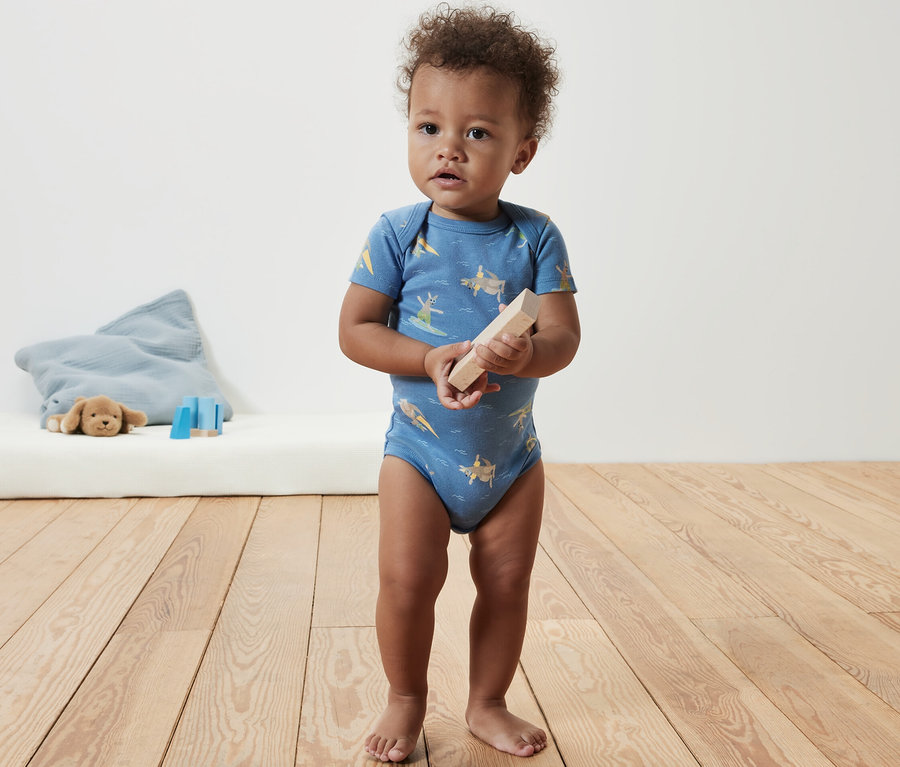 Kleinkind steht und hält einen Holzklotz, es trägt einen blauen Kurzarmbody.