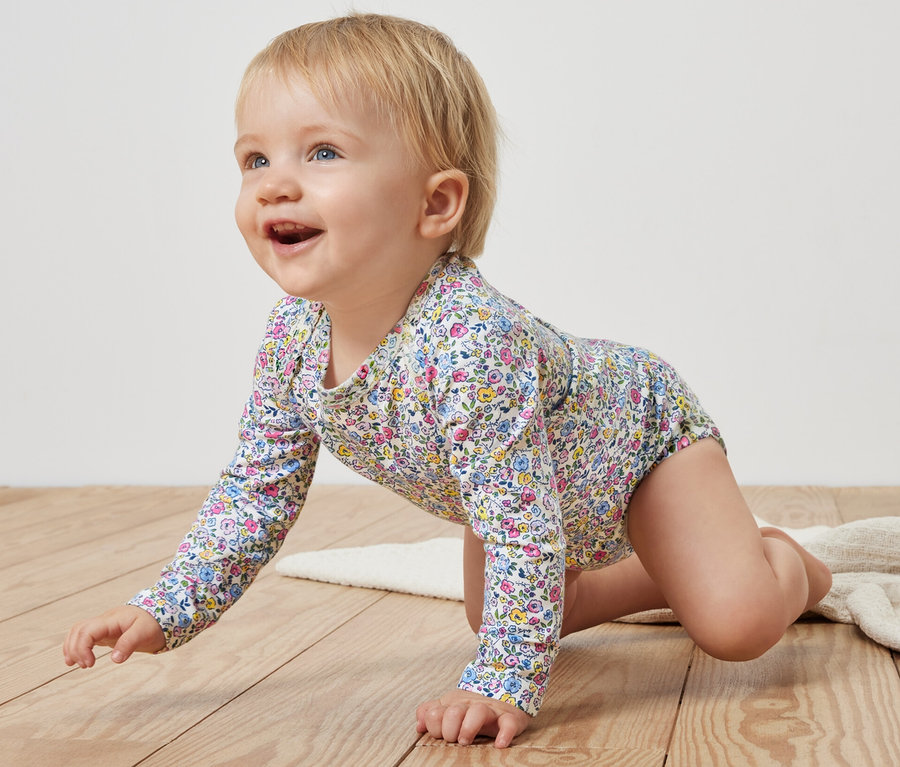 Ein Baby krabbelt auf einem Holzboden und trägt einen rosa Baby-Body mit Blumenmuster.