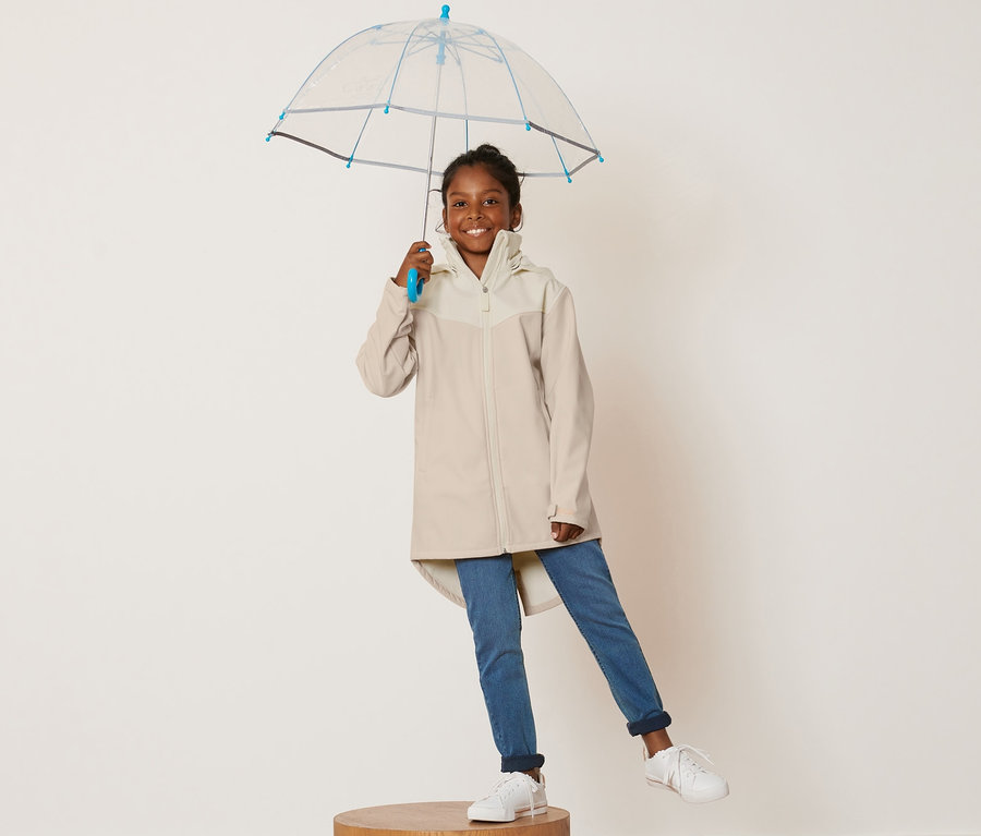 Ein Mädchen steht auf einem Holzsockel und hält einen durchsichtigen Regenschirm. Sie trägt einen Kinder-Softshellmantel.