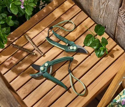 Zwei Gartenscheren liegen neben Blättern und Ästen auf einem Holztisch.