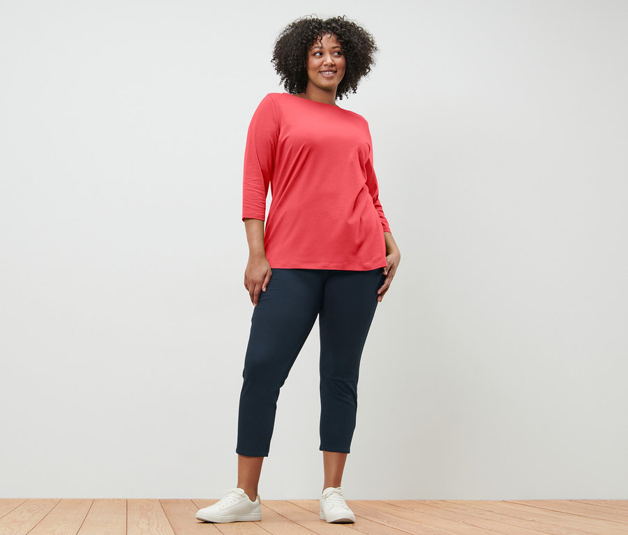 Frau posiert in einem korallenfarbenen Shirt mit 3/4-Arm und dunkelblauen Hosen.