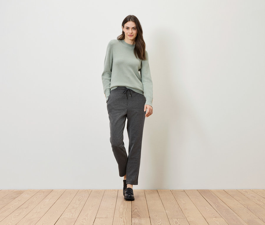 Eine Frau steht auf einem Holzboden vor einer weißen Wand. Sie trägt einen grünen Pullover und graue Hosen.
