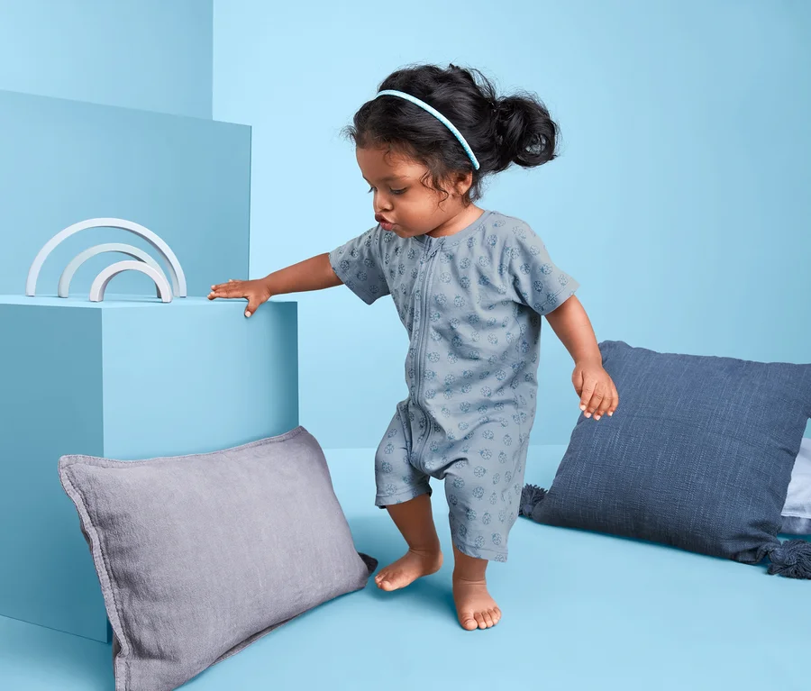 Kleinkind in grün-blauem 2er-Shorty-Pyjama steht neben Kissen und Spielzeug.