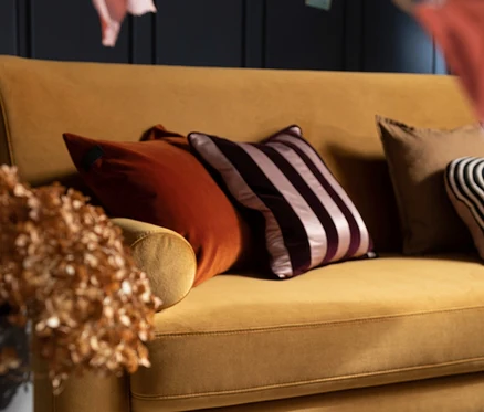 Senfgelbes Sofa mit einem terrafarbenen SCHÖNER WOHNEN-Kollektion Dekokissenbezug »SOUL«, einem gestreiften Kissen und weiteren Zierkissen. Getrocknete Blumen in einer Vase im Vordergrund.